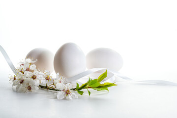 Three white Easter eggs with a small branch of wild fruit flowers on a light background with copy space, selected focus, narrow depth of field
