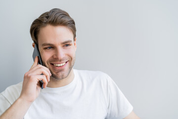 happy young man in white t-shirt talking on smartphone isolated on grey.