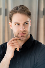 pensive man in black shirt holding hand near face and looking away at home.