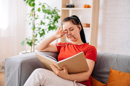 A tired young woman taking off her glasses massages her eyes after reading a book. the feeling of discomfort due to prolonged wearing of glasses, the concept of pain in the eyes. myopia