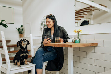 An optimistic woman in a gray suit smiles and plays with a dog in a cafe. Pretty woman in stylish coat posing