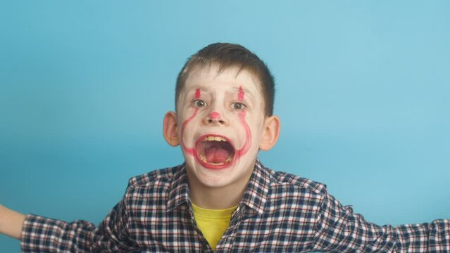 Caucasian boy 7-8 years old in scary clown makeup hides behind balloons scares looking at the camera. Studio shooting blue background. Creepy child for Halloween