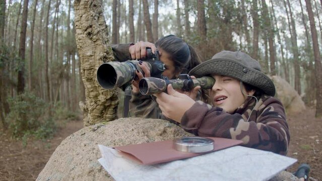 Active mother and son dressed in camouflage clothes photo hunting in forest, hiding behind stone. Happy woman taking wildlife photos, then showing shot to boy with spy pipe. Nature, leisure concept