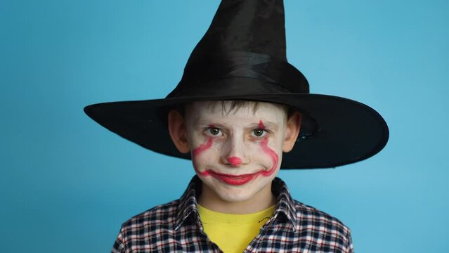 Caucasian peppy child 8 years old painted face with makeup in a hat for the Halloween holiday.Child scares with a painted clown makeup face. Horror movie. studio shooting