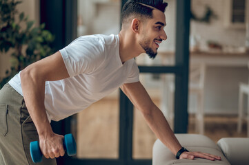 Fototapeta premium Young dark-haired man exercising with dumbbells and looking contented