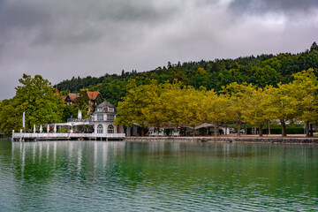 Obraz premium Mirrored calm lake water, white house and green trees line, cloudy blue sky in an autumn morning