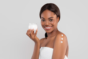 Smiling pretty millennial african american woman with perfect skin in towel with cream on shoulder showing jar