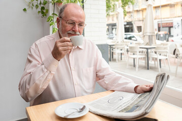 Fototapeta na wymiar a smiling older retired man with glasses reading a daily newspaper sitting in a coffee shop while drinking his coffee
