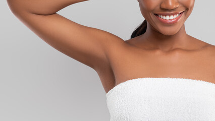 Happy millennial african american female with perfect skin in towel showing armpit isolated on gray...