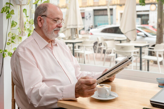an elderly retired man in glasses reading a daily newspaper while drinking his coffee, concept leisure and free time, media