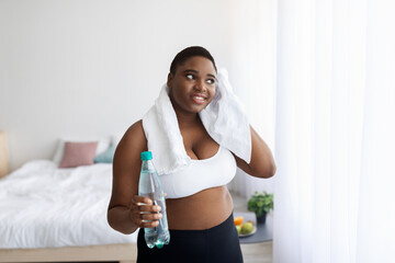 Fototapeta na wymiar Plus size young African American woman with bottle of water wiping sweat with towel after home workout