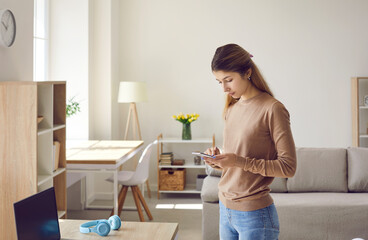 Woman using mobile phone at home. Serious young lady standing at work desk with notebook PC, holding cellphone, searching for information, messaging on social media and answering text on messenger app