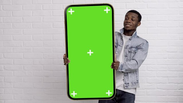 Excited black man standing and pointing Big smartphone with blank green screen, demonstrating copy space for app or website design, standing over white brick background, mockup image