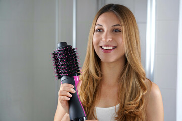 Beatiful girl holds round brush hair dryer to style hair at home. Young woman showing salon...