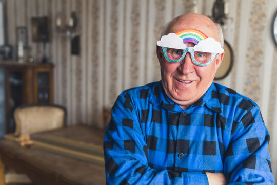 Old european bald man in a blue flannel shirt and rainbow glasses looking at camera and standing in his vintage living room. High quality photo