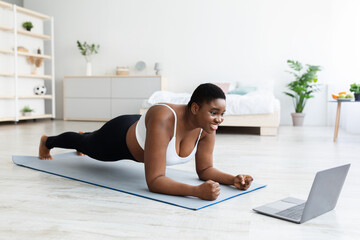 Fototapeta na wymiar Online sports and weight loss. Overweight young black woman doing exercises in front of laptop at home, full length