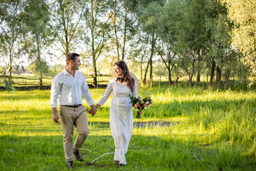 Naklejka na ściany i meble The couple walks across the field. They holds hans. The veil dissipates. In the hands of a bouquet.