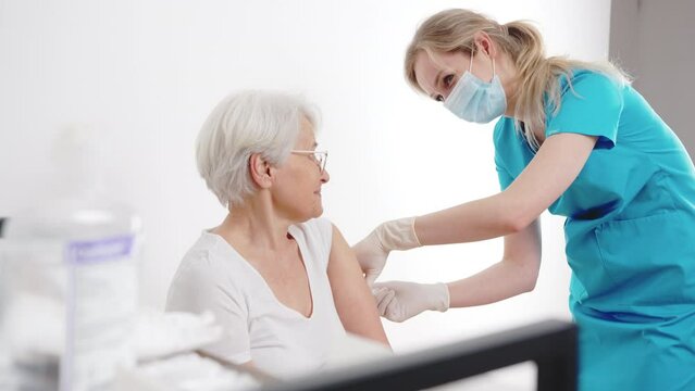 Young attractive blonde nurse vaccinating the senior gray haired lady in hospital room, immunization against virus infections. High quality 4k footage
