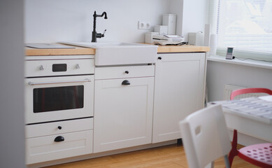 The interior of a kitchen in white tones with a sink, sandwich maker, stove, oven, dining table and a light window