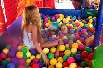 Fototapeta na wymiar The older sister plays with her younger brother and throws small balls at him while sitting in the playroom