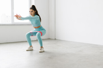 Slim woman works out with resistance band on white wall background indoor. Sporty female in...