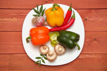 Red pepper. vegetable concept on a white plate. Two red hot peppers, a long green cucumber, a head of garlic, champignons, mushrooms, onions. on a wooden background.