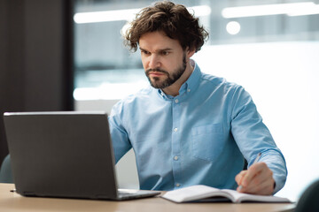 Portrait of focused man using laptop and writing in notebook