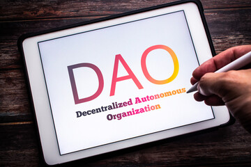 The sign of DAO (Decentralized Autonomous Organization),  an organization represented by rules encoded as a transparent computer program, on tablet on wooden table. Man hand holding wireless stylus - obrazy, fototapety, plakaty
