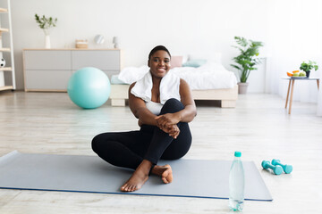 Smiling overweight young black woman sitting on yoga mat, resting after home fitness, copy space