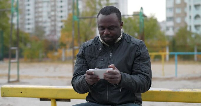African american man sits at sport ground plays the game on his smartphone. Cinema 4K 60fps video