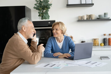 Elderly married couple laughing, talking on smartphone, sitting on kitchen