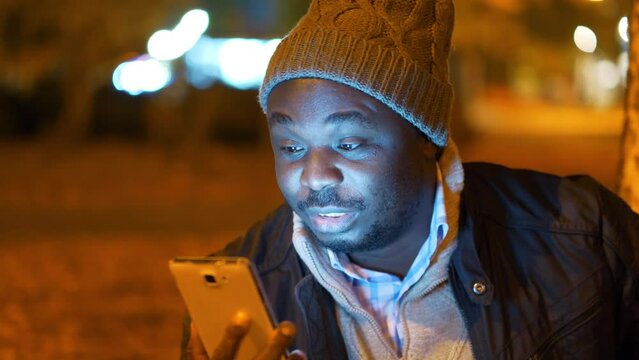 African american man in winter hat sits in evening city park and speaks via smartphone by video link