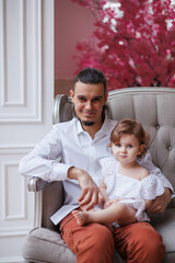 Obraz premium Handsome happy father sitting with his little daughter on beige sofa indoors. Young man in white shirt with his 1-year-old baby daughter in white dress, holding her in hands on legs. family tenderness