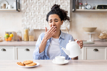 Fototapeta na wymiar Sleepy tired woman waking up early, pouring tea into cup and yawning, sitting in modern kitchen interior, copy space