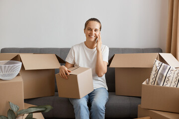 Indoor shot of attractive smiling woman wearing white shirt and jeans sitting on sofa, holding cardboard box with personal things and talking via mobile phone, looking at camera with smile.