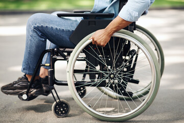 Unrecognizable young black man in wheelchair going for walk in city park, closeup of legs
