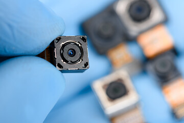Smartphone camera module in researcher hand, with other cell phone camera sensor on background. - 498783772