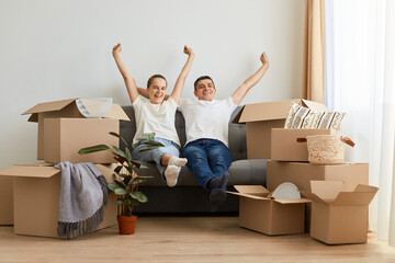 Indoor shot of young couple sitting on cough with raised hands, being happy, rejoice to move in a...