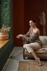 Obraz premium beautiful young woman with black hair in cozy natural clothes sits on a bright sofa near a large window