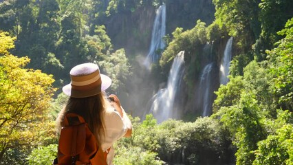 Back view of travel blogger woman on trip or adventure on Tee Lor Su waterfall in Thailand. Young female traveler make photos on smart phone for social media. Generation Z new concept travels