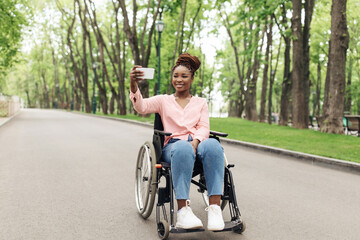 Fototapeta na wymiar Full length of happy young black woman in wheelchair taking selfie on cellphone at park, copy space