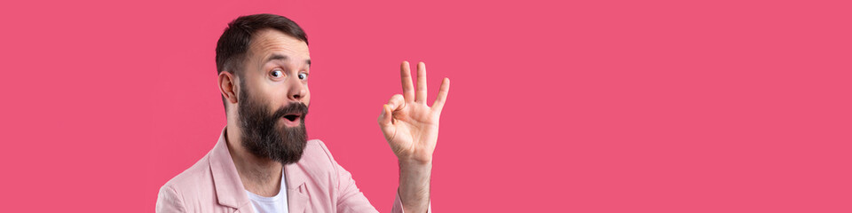 Portrait of a smiling young man in a pink jacket. Man showing ok gesture while standing in studio over isolated red background.