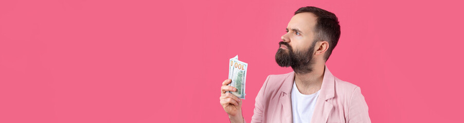 Obraz premium Portrait of a contented young businessman with a beard dressed in a pink jacket showing us dollar banknotes against a red studio background. Taste, smell of money.