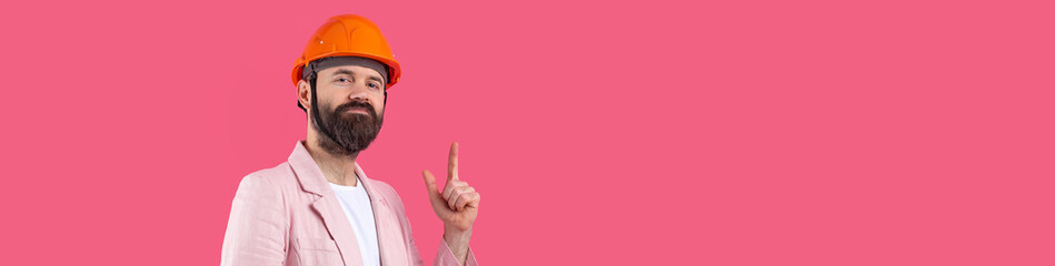 Portrait of young construction engineer wear orange hard hat, in a pink jacket standing on red studio background. The man points with his hand.