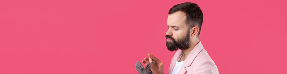 Fototapeta premium Portrait of a contented young businessman with a beard dressed in a pink jacket showing us dollar banknotes against a red studio background. Taste, smell of money.