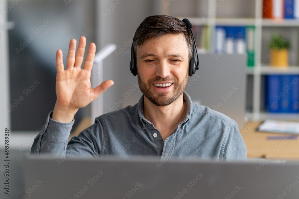 Poster Distant communication. Happy mature man in headset waving hand at computer webcamera while making video call in office