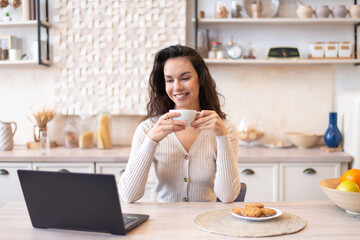 Happy woman sitting in front of laptop at kitchen table, watching movie on computer and drinking coffee