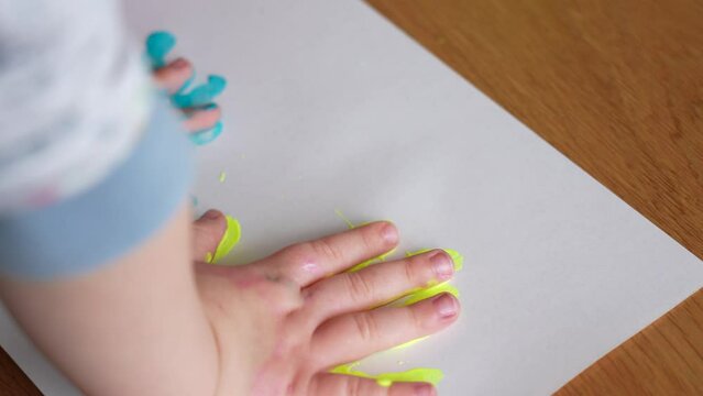 Close-up view 4k stock video footage of small hands of 4 or 5 years old baby painted in bright blue and yellow colors of national Ukrainian flag. Baby making cute handprints on white paper sheet