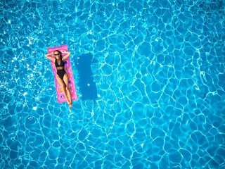 Fit pretty girl in bikini chilling on inflatable pink mattress in swimming pool. Slim hot woman in swimwear tanning. Female relaxing on float in blue water at luxury resort. Aerial, view from above.