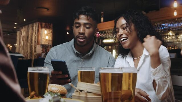 Two multiracial friends at a restaurant looking at the selfies they took on one of their mobile phones.